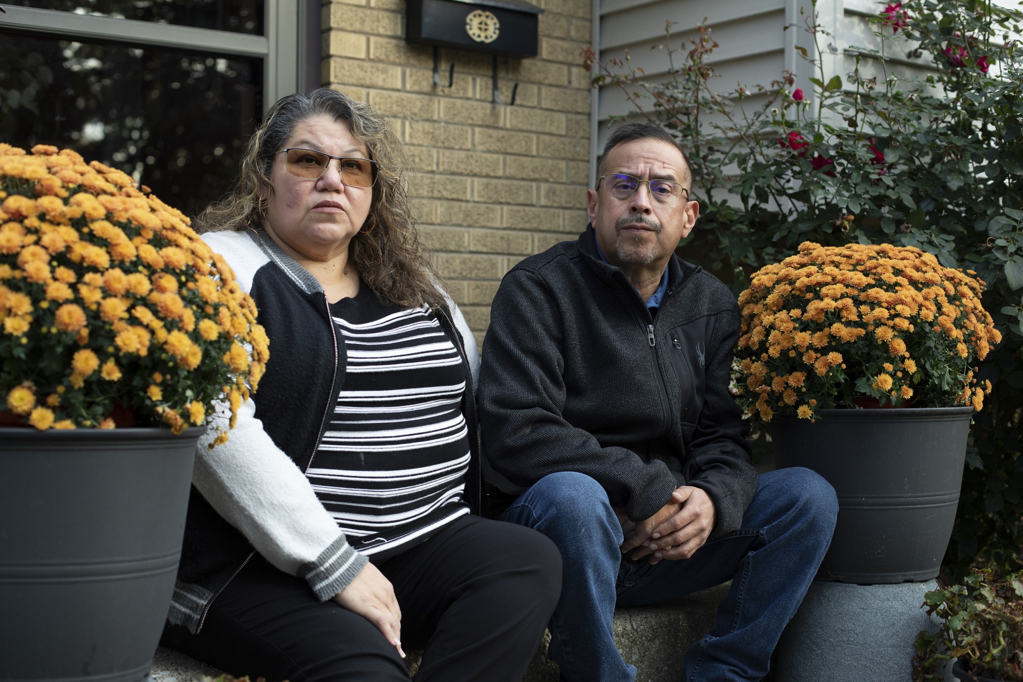 A woman and man sit on a stoop, potted marigold flowers are on either side of them. 