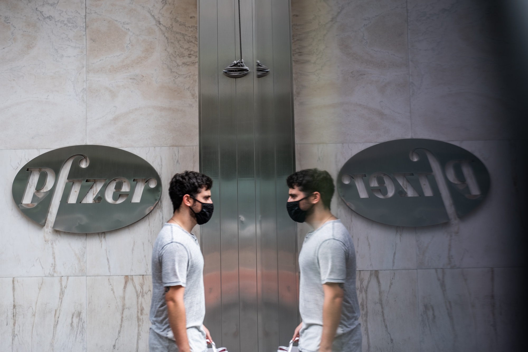 A pedestrian wearing a protective mask passing in front of Pfizer Inc. signage is reflected outside the company's headquarters in New York.
