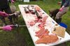 Two short-haired tawny dogs and three humans surround a white foldable table set on grass. The humans are wearing pink gloves and dark rubber boots. One of them is holding a bloody knife in the left hand and a hose in the right hand spraying water onto the table. The table has four salmon without heads on one side, blood in the middle and a pile of filets at the other end.