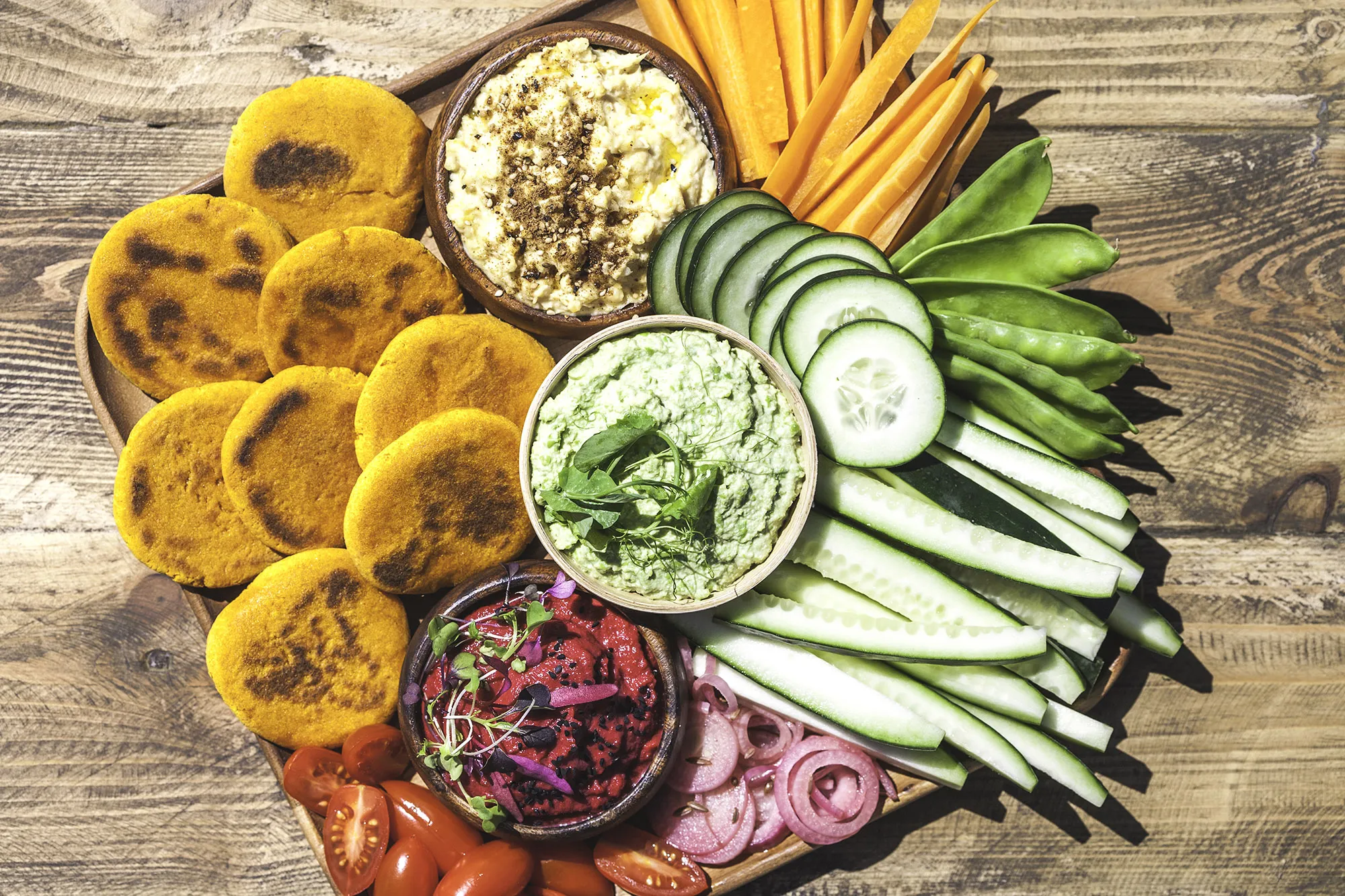 A platter of snacks for the plant-based eaters at a party. Golden turmeric flatbreads, fresh vegetable crudité and three hummus flavors; classic chickpea, spring green pea and beetroot/orange.