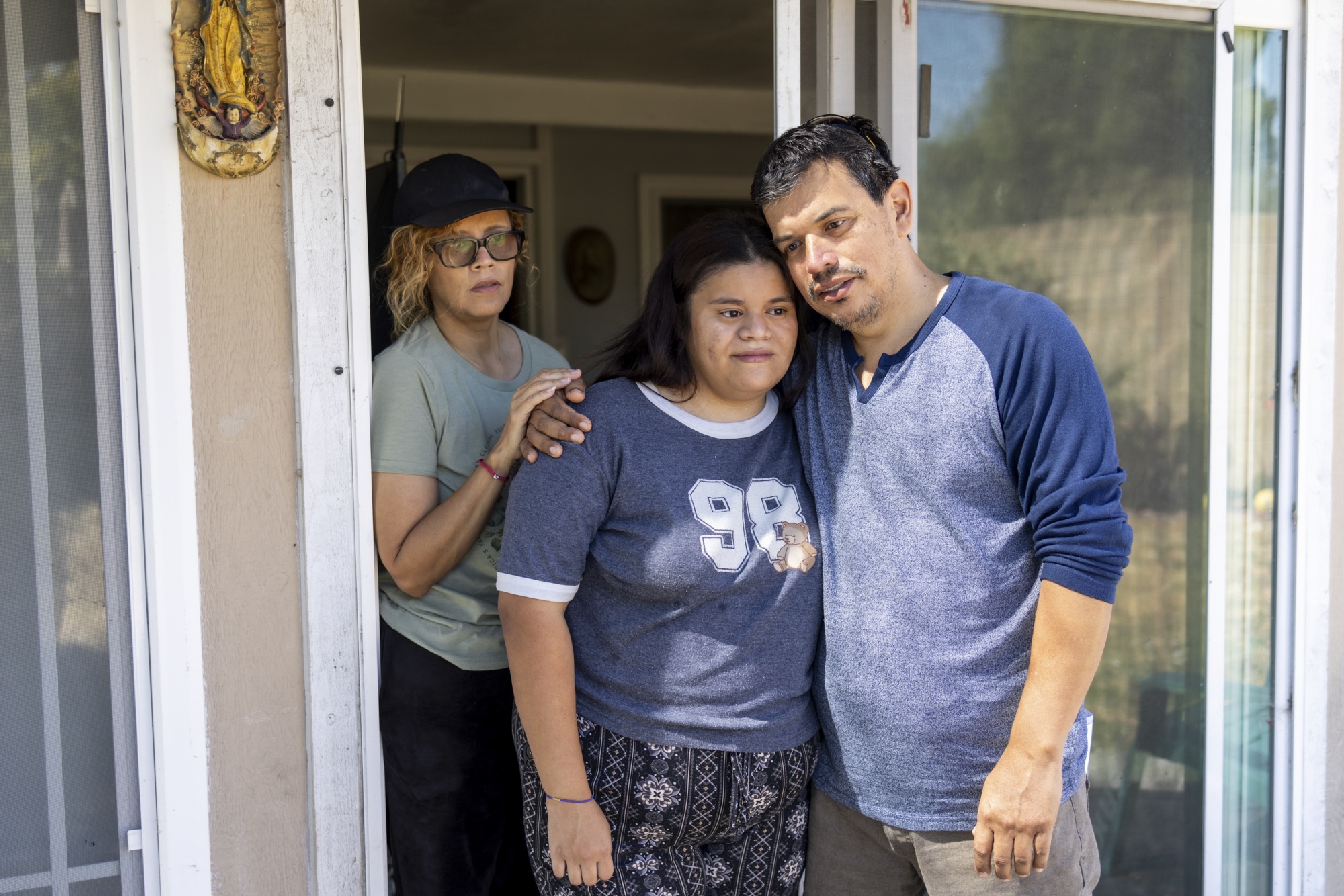 A mother places her hand on her daughter shoulder who looks into the distance. Her father places her arm around her. 