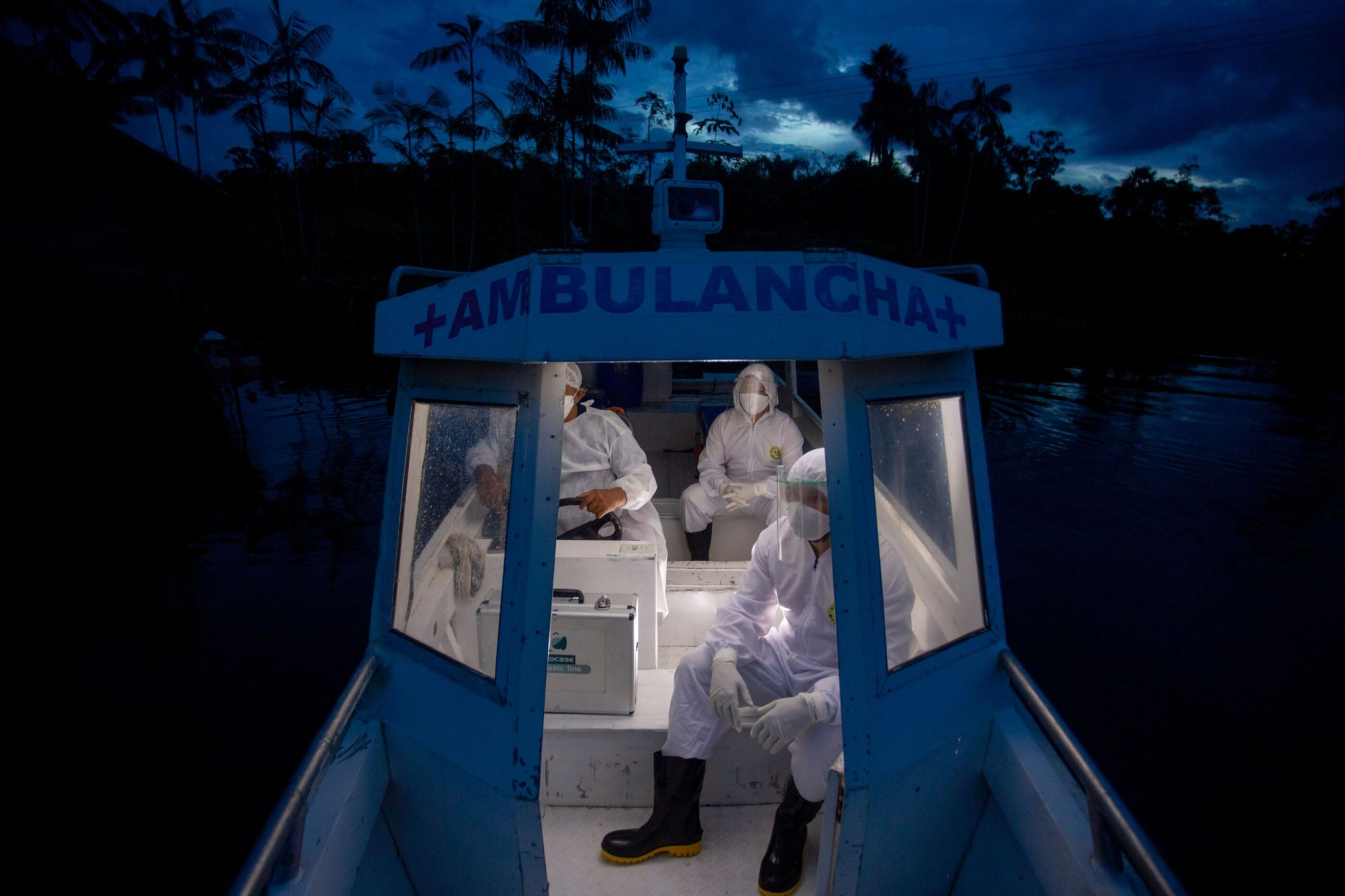 Three health workers in protective white jumpsuits, face masks and face shields ride in a small boat at night.
