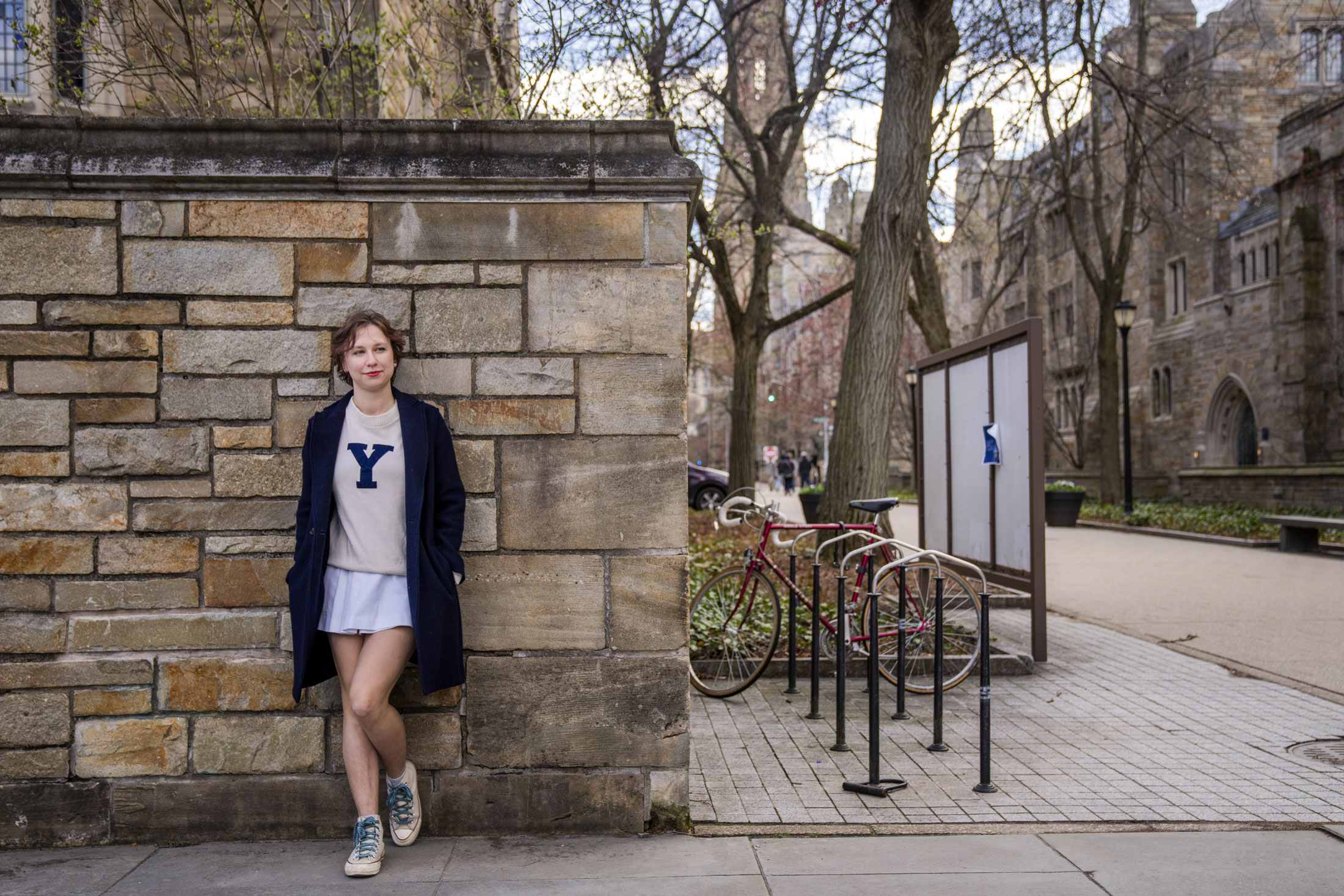 A portrait of a young woman wearing a long cardigan and shirt with a Yale Y logo, leaning against a wall