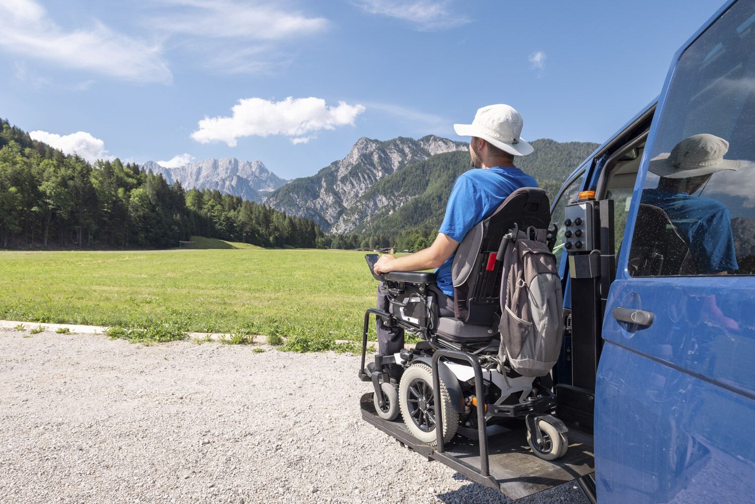 View from behind of man in a wheelchair getting out of a van with a wheelchair lift, mountains in the background. 