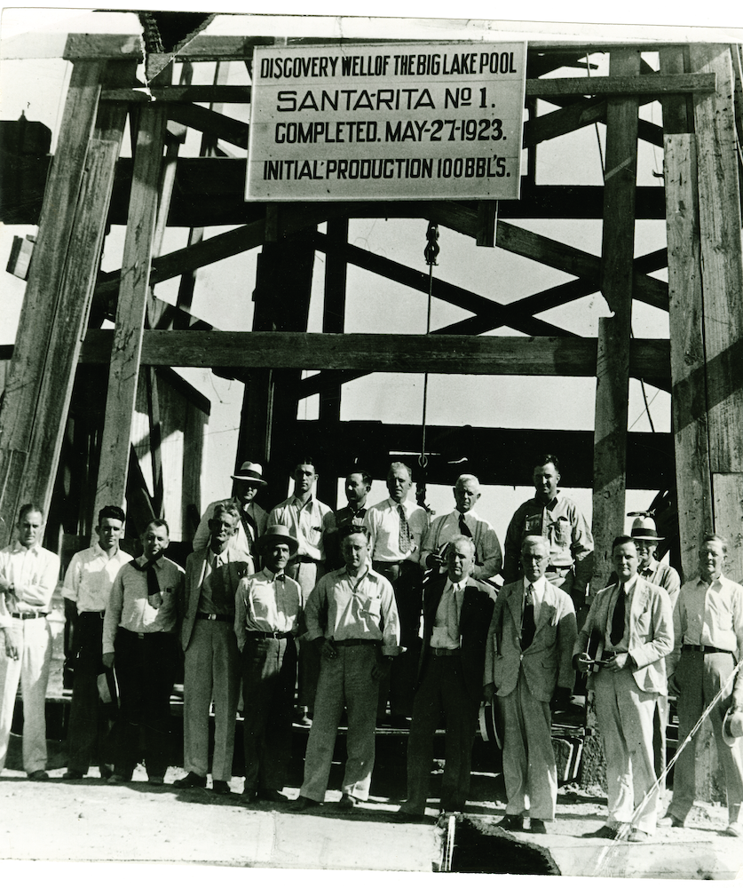 A black and white historical photo of a large group of men standing in front of the wooden well structure wearing slacks and button down shirts 