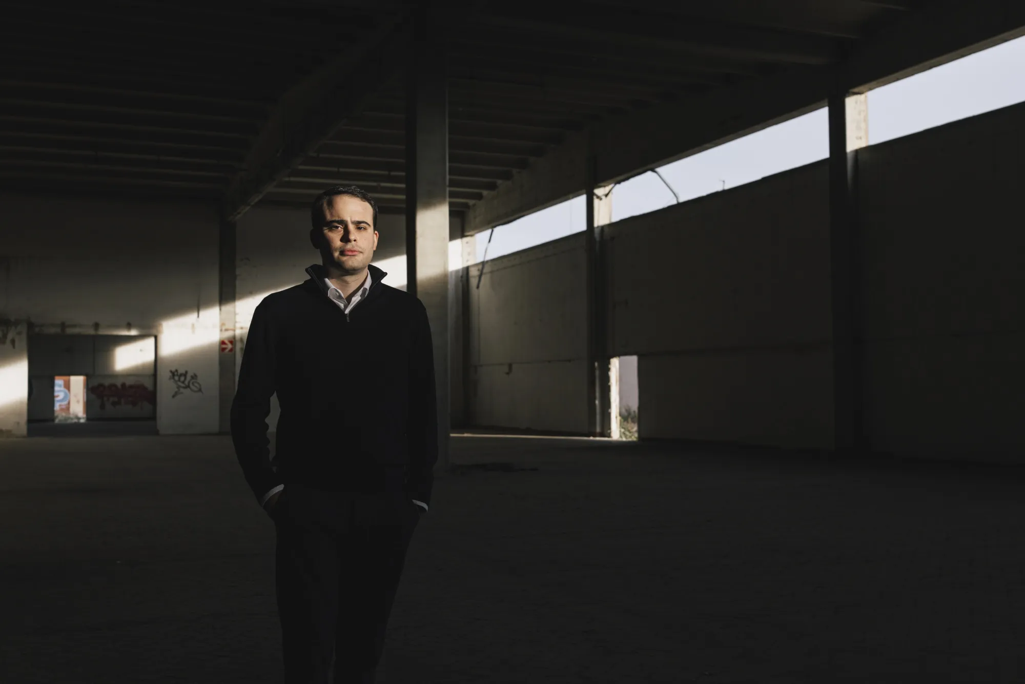 Lorenzo Avello, wearing a sweater over a white, collared shirt, stands in a large dark warehouse-like room with both hands in his pockets. Behind him the empty warehouse is sparsely lit bit narrow windows; graffiti can be seen on one wall behind Avello.