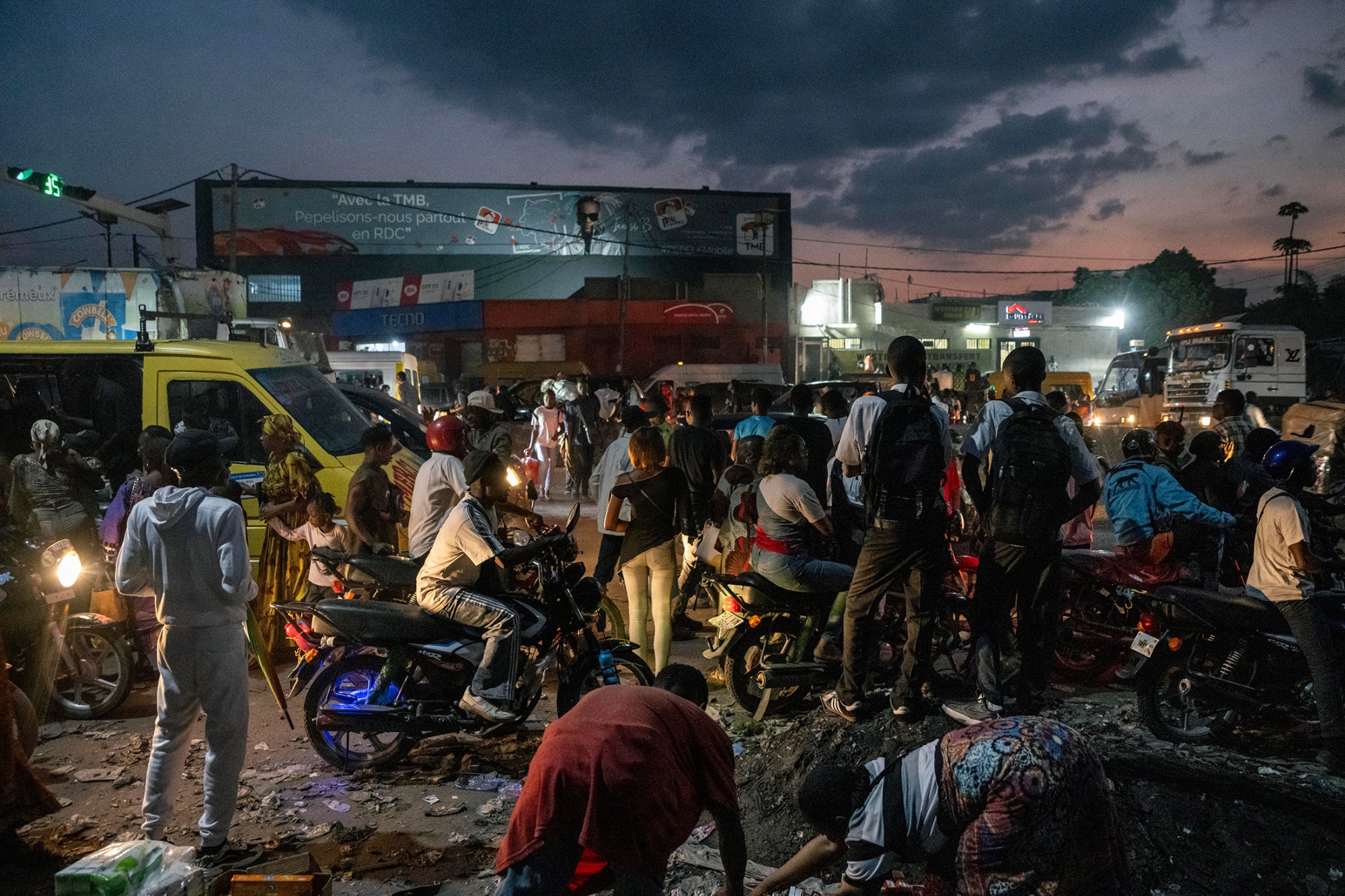 Crowded evening street scene in Kinshasa, Democratic Republic of Congo, with people, motorcycles, and vehicles gathered under dim lighting during rush hour.