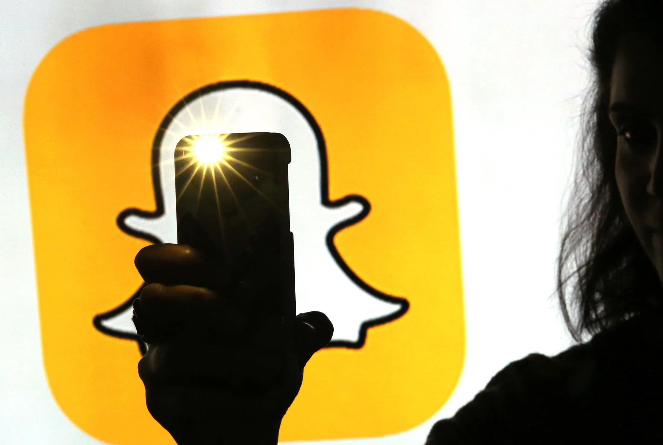 A woman takes a photograph with a camera whilst standing against an illuminated wall bearing Snapchat Inc.'s logo.