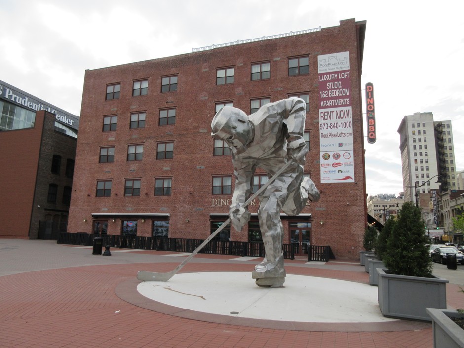 &quot;The Iron Man&quot; sculpture of a hockey player next to the Prudential Center in Newark, where the New Jersey Devils hockey team plays. In the background, signs can be seen for Dinosaur Bar-B-Cue BBQ and Rock Plaza Lofts luxury apartments.