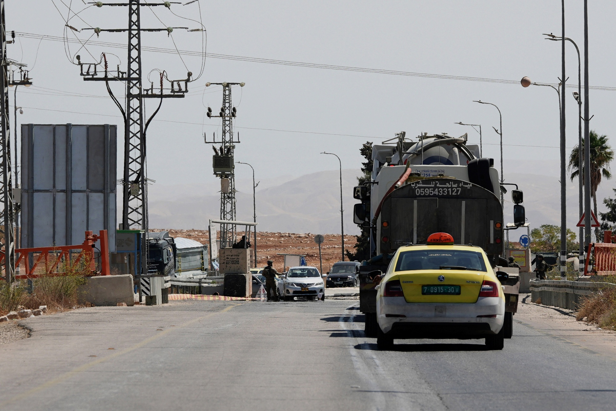 Vehicles, including a yellow taxi and a large truck, wait in line at a checkpoint outside Taybeh, with soldiers visible near the barricades and hills in the background.