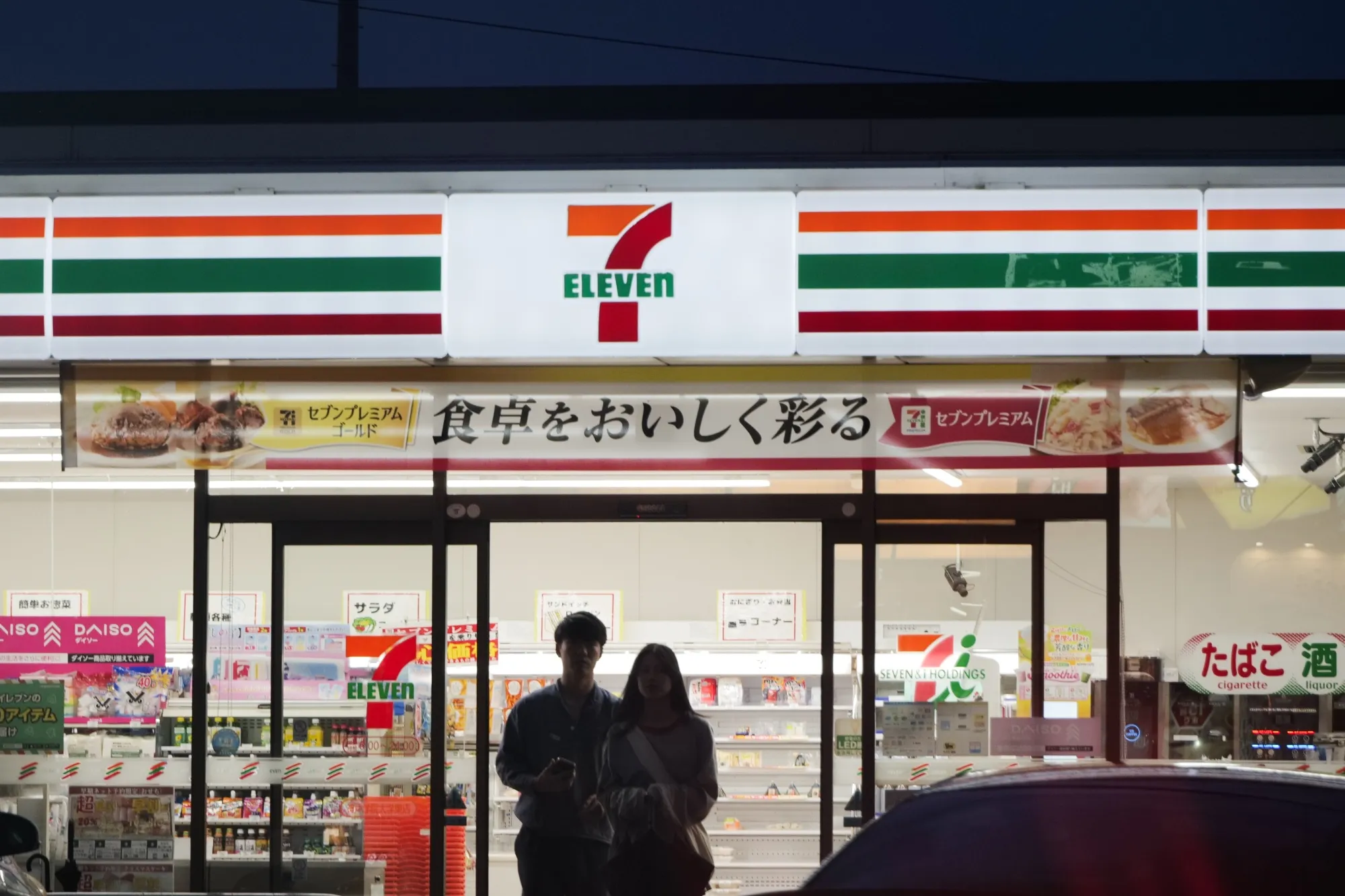 Customers exit a 7-Eleven convenience store, operated by Seven &amp; i in Kobe, Japan.