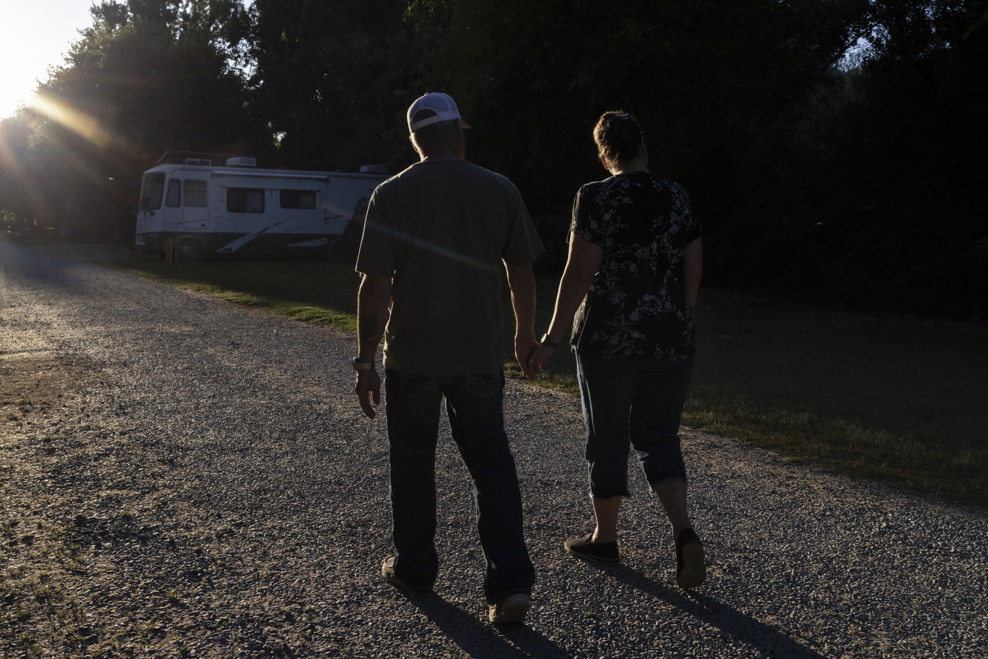 A couple walk down a gravel road holding hands while the sun sets, an RV can be seen in the background.