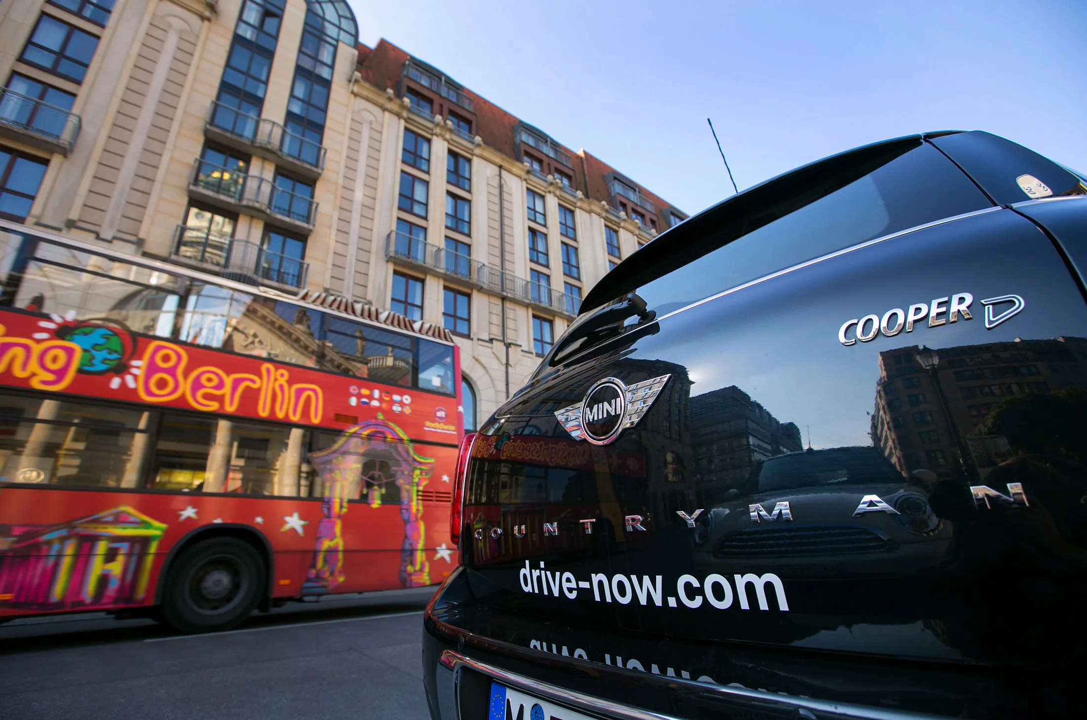 A sightseeing bus passes a BMW Mini Cooper D DriveNow car as it sits parked on a city center street in Berlin.
