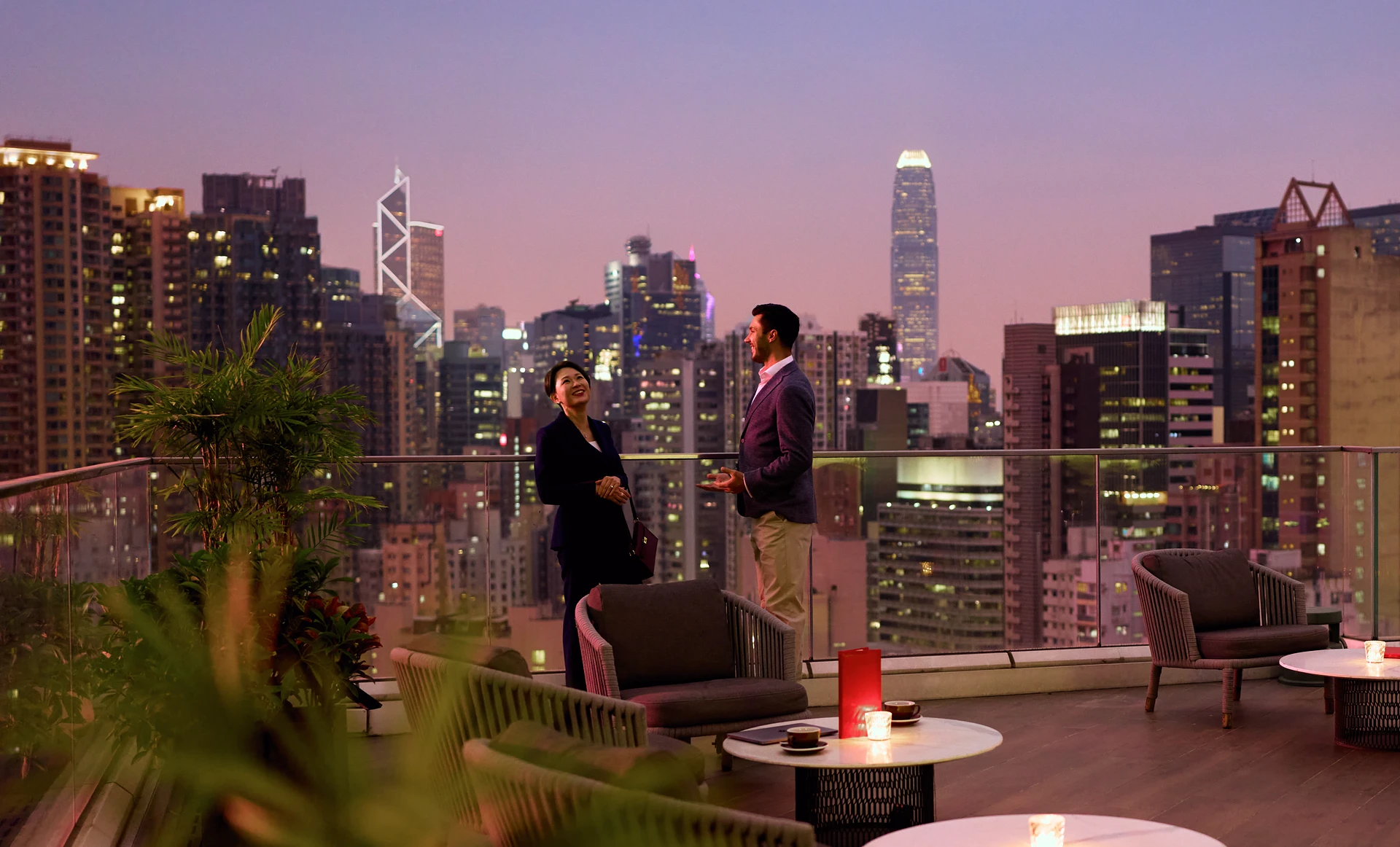 A man and a woman in smart-casual attire stand conversing on a rooftop terrace at dusk, surrounded by comfortable lounge chairs and small tables. In the background, the illuminated skyline of Hong Kong features prominent skyscrapers, including the Bank of China Tower and Two International Finance Centre, under a purple-tinted evening sky.