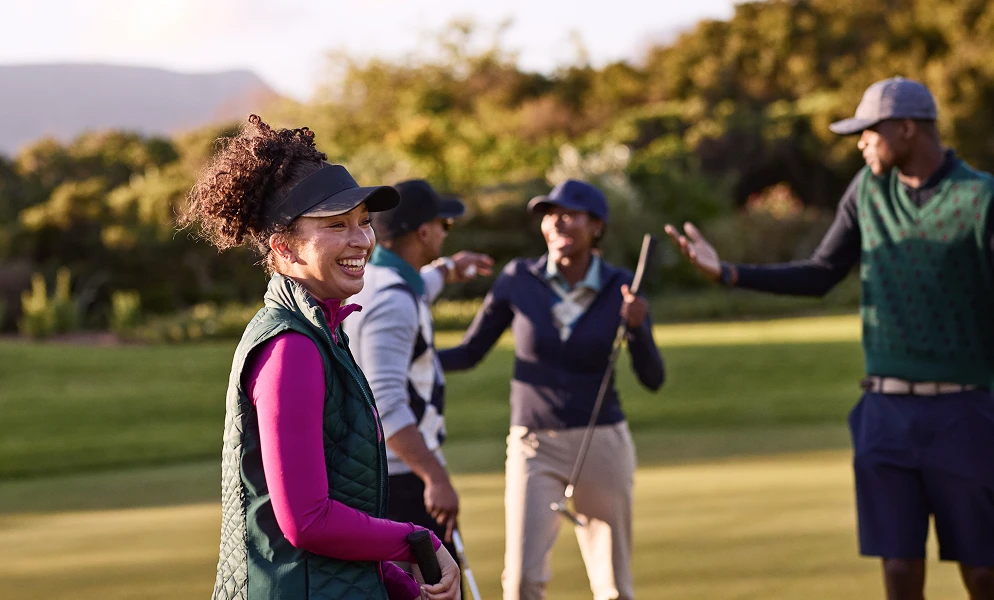 A cheerful woman with curly hair tied back in a visor smiles in the foreground, wearing a bright pink long-sleeve shirt and a dark green quilted vest on a golf course. In the background, three of her friends interact in a relaxed, friendly atmosphere while holding golf clubs under warm sunlight.