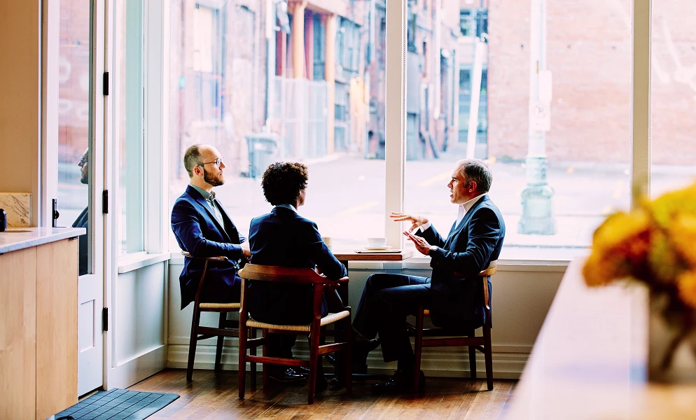 Three people dressed in business attire sit around a small wooden table next to large floor-to-ceiling windows inside a brightly lit room. A man on the right with graying hair gestures with his hands while speaking to the other two individuals. The person in the middle sits with their back toward the camera, and a man wearing glasses on the left listens attentively. Outside the window, a city street and brick buildings are visible.