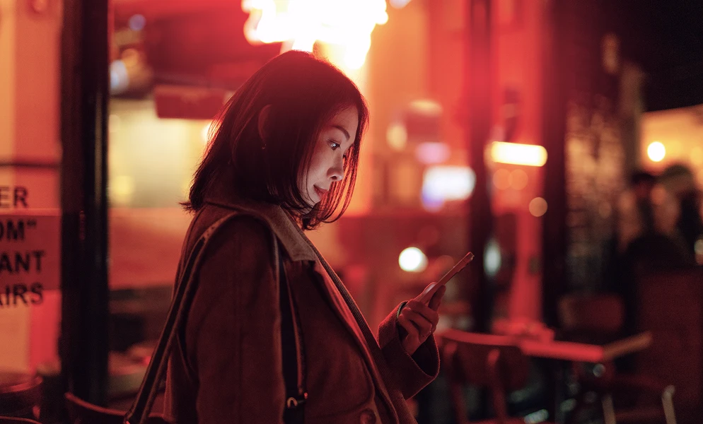 A profile view of a young woman with shoulder-length dark hair looking down at her mobile phone, illuminated by glowing red and neon lights in a night urban setting.