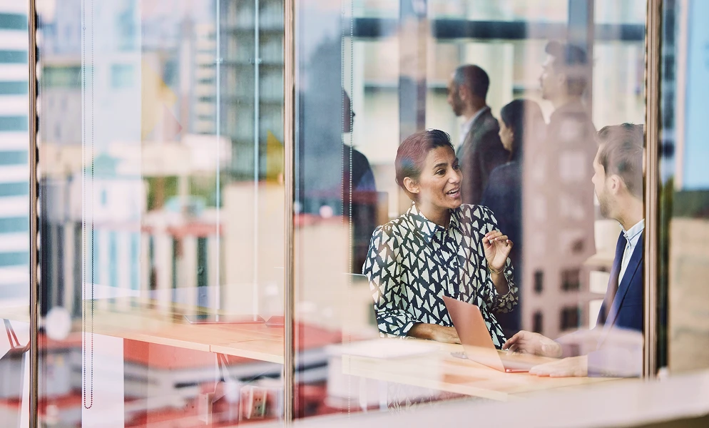 A woman wearing a patterned black-and-white shirt sits at a wooden table in an office, gesturing while conversing with a colleague in a suit. A laptop sits open on the table between them. They are viewed through a glass window wall, which reflects the surrounding cityscape and shadows of other people standing in the background.