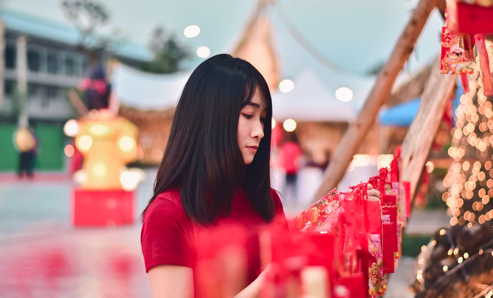 A young woman with dark, shoulder-length hair and bangs wears a red short-sleeved shirt as she looks down while adjusting hanging red decorations or envelopes. The outdoor setting features festive lights with soft bokeh effects, white tents in the background, and additional red festive displays surrounding her.