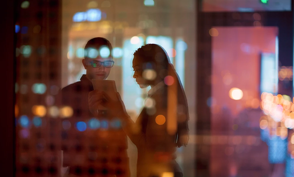 A man wearing glasses and a woman stand side-by-side indoors, looking down at a small tablet device that the man is holding. The photograph is taken from outside through a reflective glass window at night, layered with soft, glowing city lights in the background and reflections across the glass.