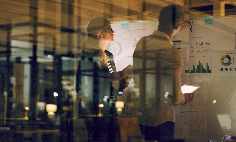 A man and a woman work late at night in an office, viewed through a glass partition. The woman stands on the left wearing glasses and a blazer, looking at a whiteboard covered with charts, graphs, and sticky notes while holding a tablet. The man, seen from behind on the right, holds a glowing tablet and writes directly on the board. Dim city or office lights reflect on the glass around them.