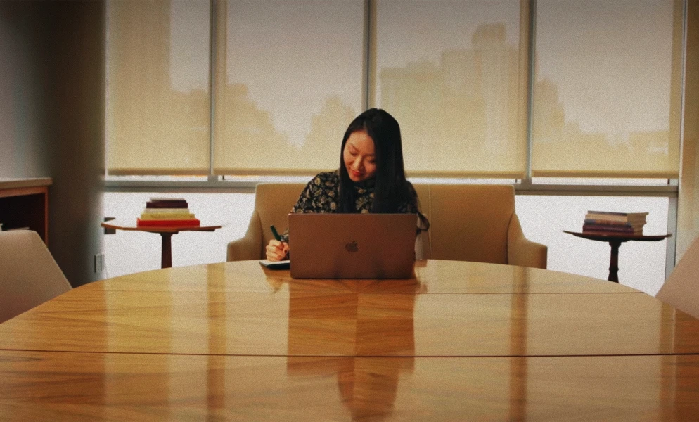 A woman with long dark hair sitting at a polished wooden oval table in front of large window blinds, using a open laptop while writing on a pad.