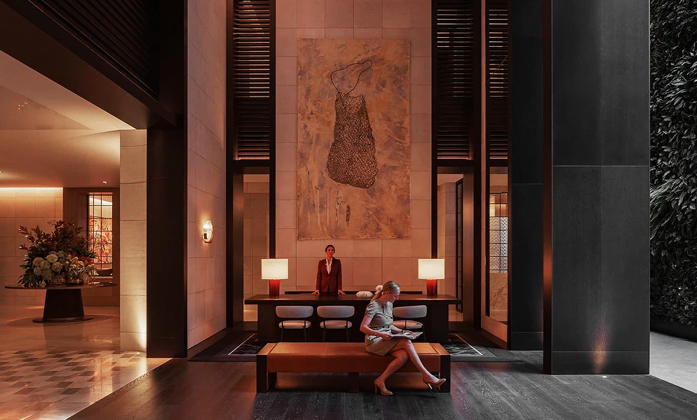 An elegant hotel lobby area with high ceilings and warm lighting. In the center, a staff member stands behind a long dark desk set against a wall with a tall abstract painting. In the foreground, a woman sits on a bench reading, while a floral arrangement is visible in an adjacent room to the left.