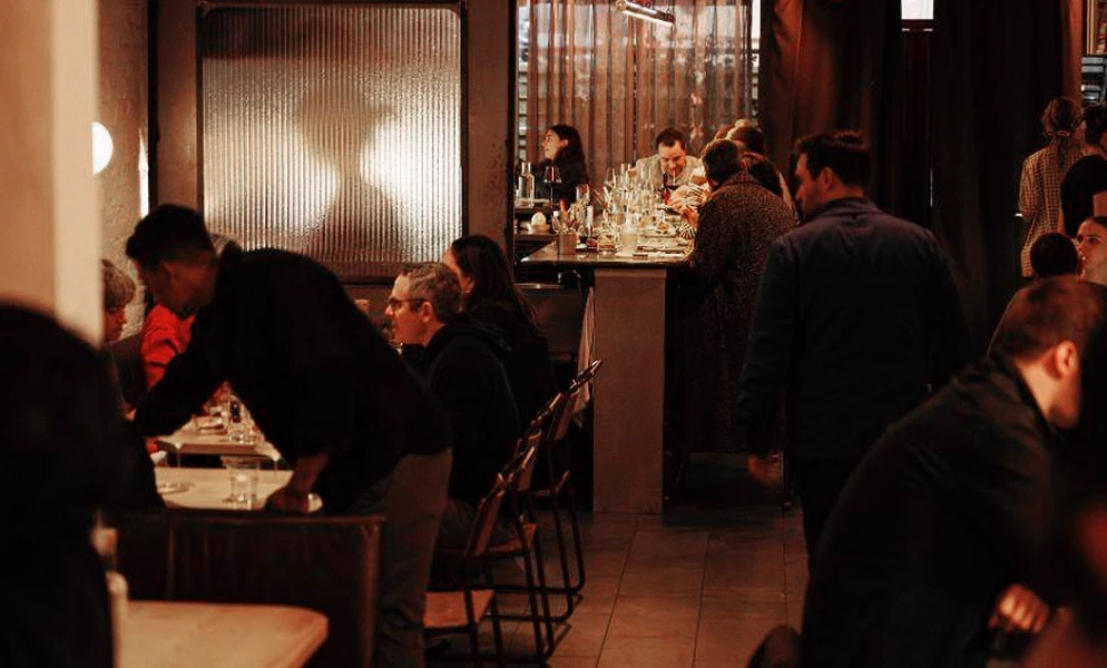 A dimly lit, bustling restaurant interior filled with patrons. In the foreground on the left, a server in a dark long-sleeved shirt bends over a table. Beside them, seated customers face toward a central high counter top where other guests are dining. On the right, a man with his back to the camera walks through the room, while another man in a dark jacket sits in the lower right corner. The atmosphere is warm with reddish ambient lighting.
