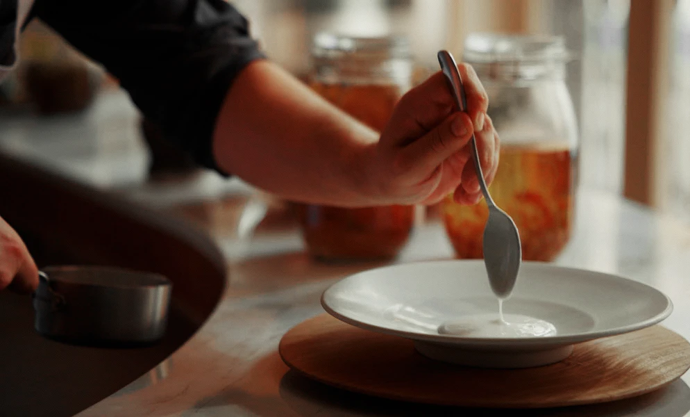 Close-up of a person using a spoon to pour a white sauce into a shallow white bowl resting on a wooden board, with glass jars in the background.