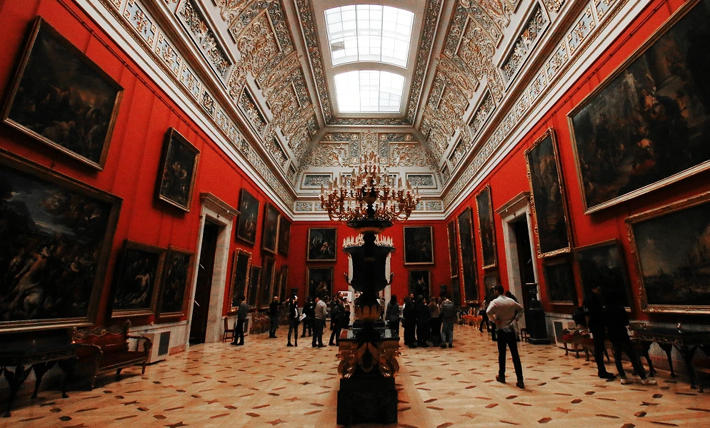 An ornate museum gallery with vibrant red walls lined with large framed paintings. The room features a vaulted ceiling with detailed gold molding and skylights, and a large chandelier hanging above a decorative pedestal in the center of the tiled floor. Several visitors are scattered throughout the hall viewing the artwork.