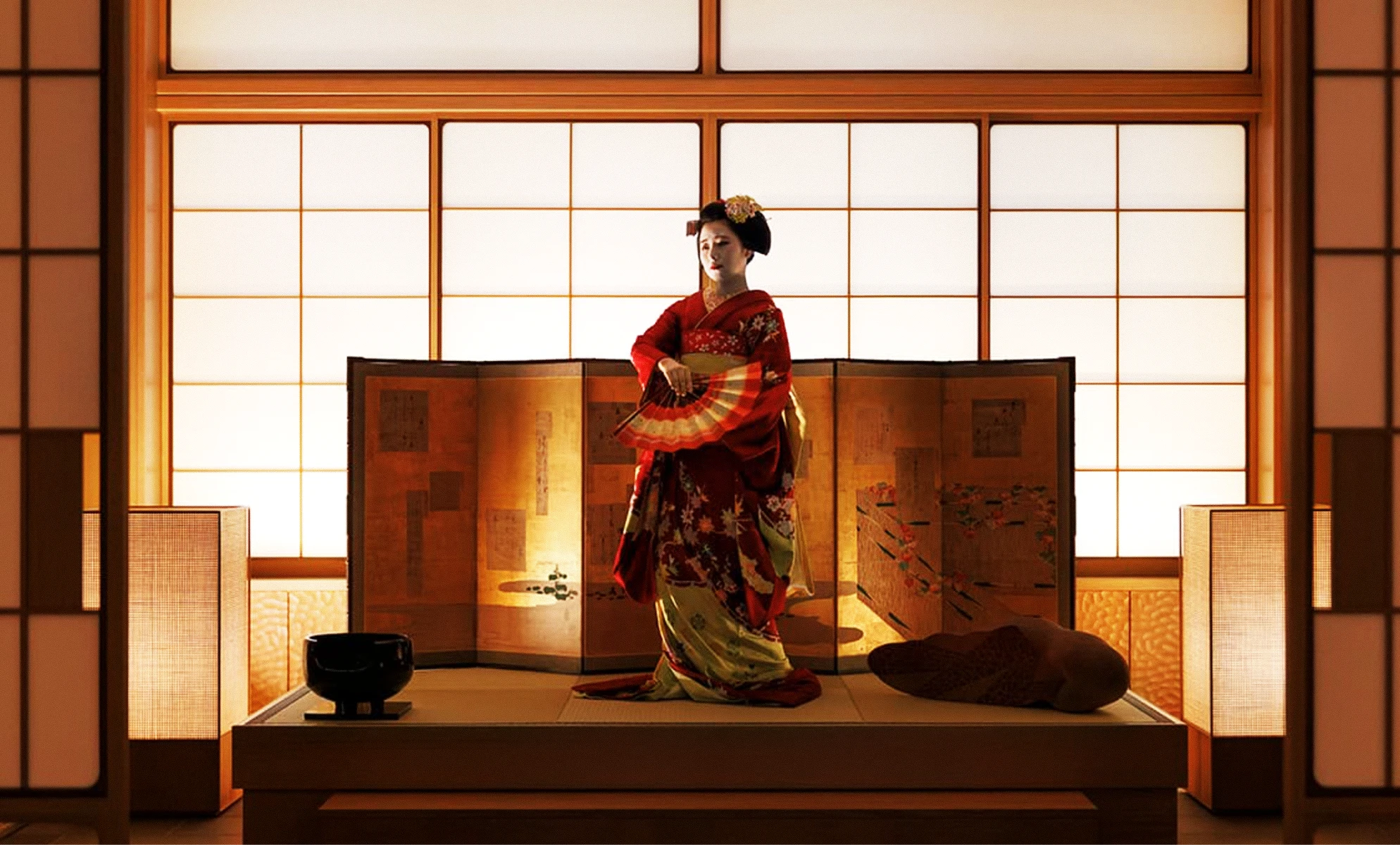 A geisha in an elaborate red and gold kimono holding an open folding fan, standing on a low wooden stage in front of a folding screen and glowing shoji panels.