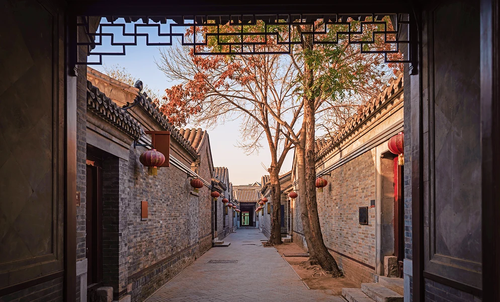 A view framed by a dark wooden doorway looking down a narrow, traditional alleyway lined with grey brick buildings topped with curved tiled roofs. Red lanterns hang along the sides of the buildings, and bare trees with autumnal leaves stand in the center.